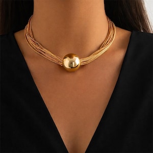 Può includere: Una collana dorata con un grande pendente sferico. La collana ha più fili di catena sottile che avvolgono il collo, con la sfera al centro. Lo sfondo è un top nero.