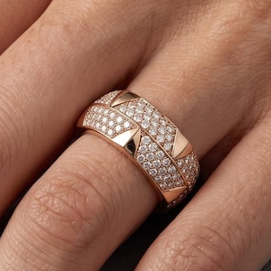 May include: Rose gold ring with a double band design. The top band is encrusted with small, sparkling diamonds, while the lower band features a row of triangular accents. The ring is displayed on a hand, showcasing its intricate details and elegant style.