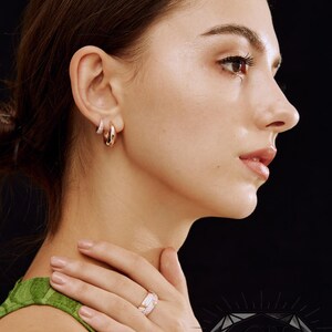 May include: A woman wearing rose gold hoop earrings and a matching ring with small, sparkling stones. The jewelry is set against a black background. The woman is wearing a green top. The ring and earrings are a matching set.