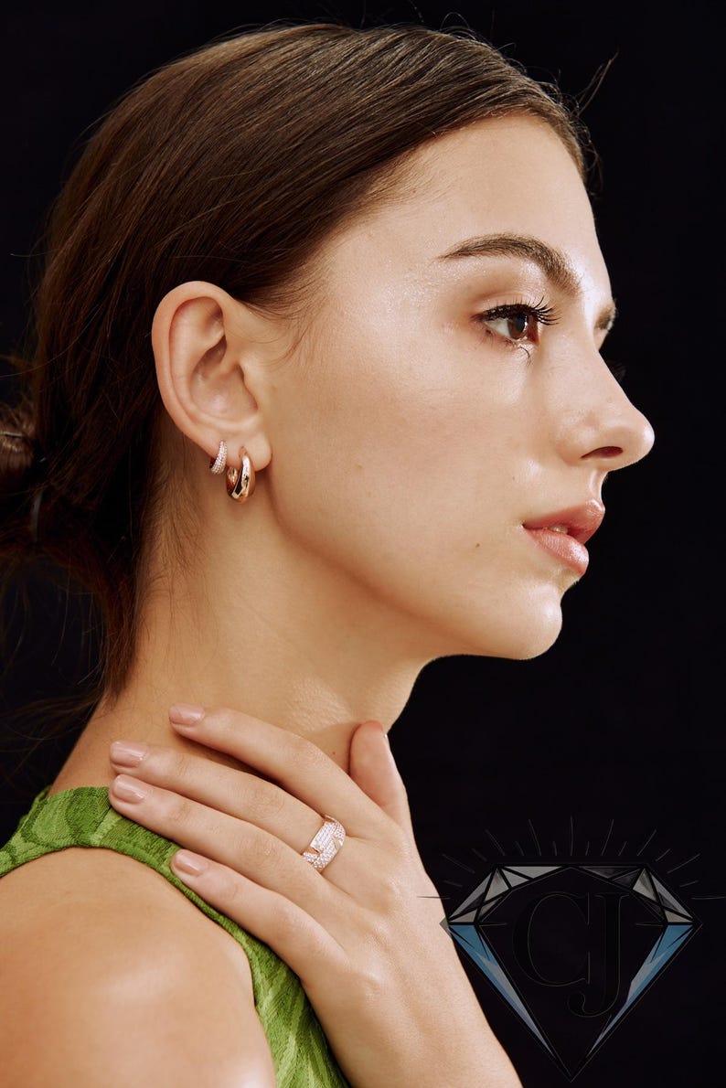 May include: A woman wearing rose gold hoop earrings and a matching ring with small, sparkling stones. The jewelry is set against a black background. The woman is wearing a green top. The ring and earrings are a matching set.