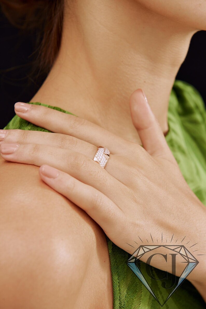 May include: A rose gold ring with a geometric design, set with small, sparkling white gemstones. The ring is on a person's finger, with the hand resting on their shoulder. The background is a soft green.