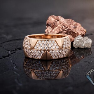 May include: A rose gold ring, heavily set with small diamonds, reflecting light. The ring has a geometric design and sits on a dark, glossy surface. A rough, reddish-brown stone and a cluster of clear crystals are in the background.