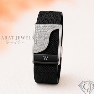 Puede incluir: Una pulsera negra texturizada con una pieza central rectangular de tono plateado que presenta una sección curva de pequeñas piedras transparentes y una barra horizontal negra grabada con la letra W. El texto CARAT JEWELS Gems of Grace es visible.