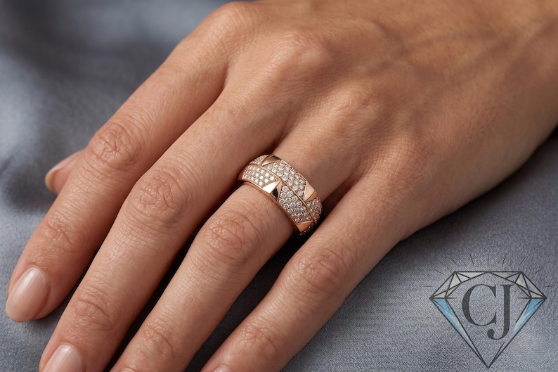 May include: Rose gold ring with a double band design. The top band is encrusted with small, sparkling diamonds, while the lower band features a row of triangular accents. The ring is displayed on a hand, showcasing its intricate details and elegant style.