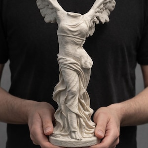 Puede incluir: Una estatua color crema de la Victoria de Samotracia, una figura drapeada sin cabeza con alas extendidas. La escultura se sostiene frente a un fondo oscuro.