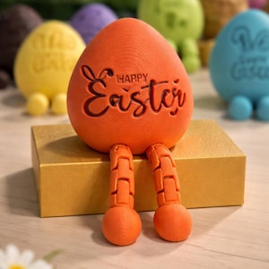 May include: An orange Easter egg ornament with the words "Happy Easter" carved into the front. The egg has orange legs and is sitting on a gold box. Other colourful Easter eggs are in the background.