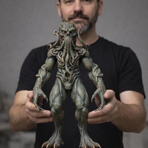 Op de afbeelding: Gedetailleerde sculptuur van een humanoïde wezen met een tentakelachtige kop, gespierd lichaam en klauwachtige handen en voeten. De figuur is voornamelijk in groen- en grijstinten, met ingewikkelde details. De sculptuur wordt voor een persoon gehouden.