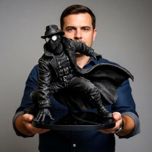 May include: A black figurine of a superhero in a trench coat, hat, and goggles, posed in a crouching position. The figure is on a black circular base. The figure is held by a person wearing a blue denim shirt.