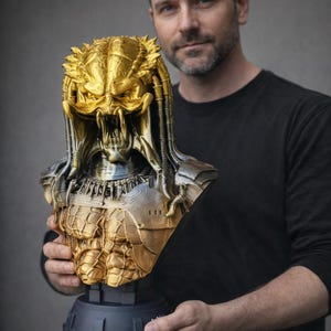 Puede incluir: Un busto de Predator dorado y plateado. La detallada escultura presenta el casco, las mandíbulas y la armadura del icónico alienígena. El busto está montado sobre una base gris oscuro, mostrando el intrincado diseño del personaje de ciencia ficción.