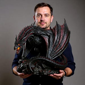 以下が含まれることがあります： 赤色のアクセントが付いた黒いドラゴンの詳細な彫刻。翼ととげが含まれます。ドラゴンはオレンジ色の目を輝かせ、不気味な表情をしています。彫刻は、灰色の背景に対して人が持っています。