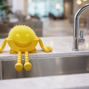 May include: A yellow sponge with a smiling face, arms, and legs, designed to sit on the edge of a sink. The sponge is approximately 10 cm in diameter. The sink is stainless steel, and the faucet is chrome.