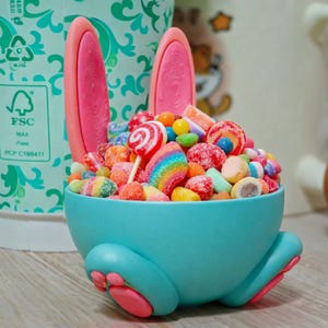 Op de afbeelding: Een turquoise snoepschaal in de vorm van een konijn, gevuld met een kleurrijk assortiment snoepjes, lolly's en gummy's. De schaal heeft roze konijnenoren en poten, wat zorgt voor een speels en feestelijk ontwerp.