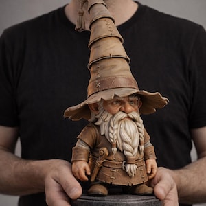 May include: A detailed figurine of a wizard gnome, featuring a long white beard and a tall, brown hat. The gnome is dressed in brown clothing and stands on a gray stone base. The figurine is held in a person's hands.