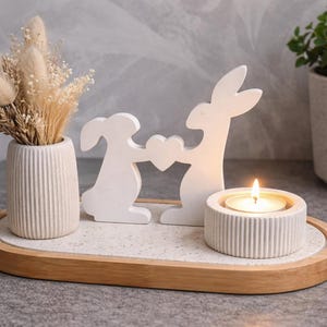Puede incluir: Centro de mesa decorativo con temática de Pascua. Incluye un jarrón blanco con flores secas, dos figuras de conejos blancos sosteniendo un corazón y una vela encendida en un soporte redondo. Los elementos están dispuestos en una bandeja de madera.