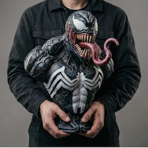 May include: A detailed bust of the Marvel Comics character Venom, held by a person. The bust features a black and white color scheme with a large, red tongue protruding from the mouth. The chest area has a white spider-like emblem.