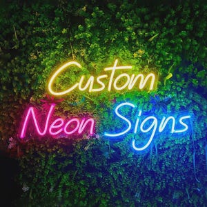 以下が含まれることがあります： 筆記体で「Custom Neon Signs」と書かれたネオンサイン。Customの文字は黄色、Neon Signsの文字はそれぞれピンクと青に発光しており、背景には密集した緑の葉が配置されています。