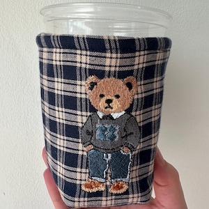 Peut inclure: Manchon de tasse à carreaux bleu marine et beige avec un motif d'ours en peluche brodé. L'ours porte un pull gris, un jean bleu et des chaussures marron. Le manchon est conçu pour une tasse en plastique transparente.