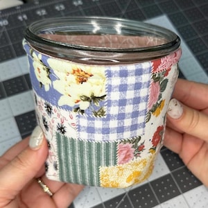 Puede incluir: Un tarro de cristal envuelto en una funda de tela de retazos. La tela presenta estampados florales, de cuadros y a rayas en tonos azules, rosas, amarillos y verdes. El tarro está sujeto por dos manos.
