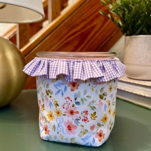 May include: A decorative jar with a floral fabric exterior featuring pink, yellow, and orange flowers with blue leaves. The jar has a ruffled, purple and white gingham trim around the rim.