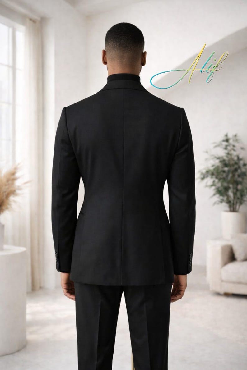 May include: A black suit, viewed from the back, with a matching blazer and trousers. The suit is tailored and has a classic design. The person is wearing a black turtleneck. The background is a bright, modern interior.