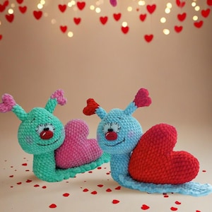 May include: Two crocheted snail toys with heart-shaped shells. One snail is teal with a pink heart, the other is blue with a red heart. Both have smiling faces and heart-shaped antennae. Red heart confetti is scattered around.