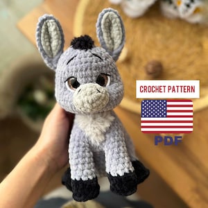 May include: A handmade gray crochet donkey with black hooves and a white chest. The donkey has large, expressive eyes and a small black tuft of hair. The image includes text that says "CROCHET PATTERN" and "PDF" with an American flag.