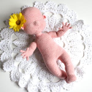 May include: A handmade, pink crocheted baby doll with closed eyes and outstretched arms and legs. A yellow flower is tucked behind one ear. The doll is lying on a white, lacy, crocheted doily.