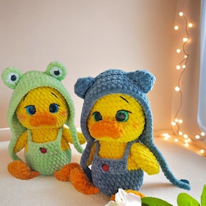 May include: Two handmade crochet duck plushies. One duck is wearing a green frog hat and overalls, the other a blue bear hat and overalls. Both have yellow bodies, orange feet, and large eyes. A red ladybug is on the overalls.