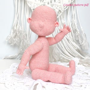 May include: A crocheted doll in a seated position, crafted in a soft pink yarn. The doll features a rounded head, small ears, and outstretched arms. The text "Crochet pattern pdf" is visible in the upper right corner. The doll is sitting on a white, lacy surface.