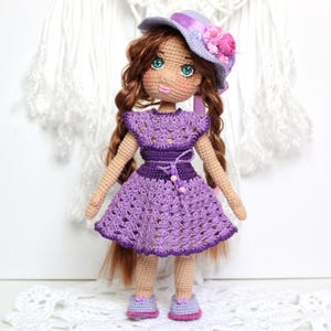 May include: A handmade crochet doll wearing a purple dress, matching shoes, and a hat adorned with flowers. The doll has long brown hair and green eyes. The dress features a detailed openwork pattern and a belt.