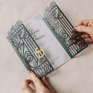 May include: A wedding invitation with a dark teal laser-cut gate design. The invitation features the names "Alexis Brian" and event details. A gold-colored number 6 is attached to the gate. The invitation is held open by two hands.