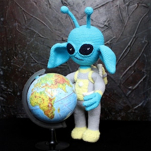 Puede incluir: Muñeco alienígena azul tejido a ganchillo con orejas grandes y ojos negros, con un mono gris y amarillo, sosteniendo un globo terráqueo. El muñeco tiene una mochila y antenas en la cabeza. El globo está sobre un soporte negro.