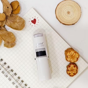 May include: A white self-inking stamp with the text "trodat" and "4612" printed on the side. The stamp is on a grid-lined notebook next to a red heart illustration and dried orange slices. A wooden slice and dried leaves are also in the image.