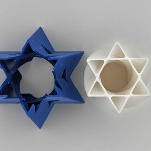 May include: Two Star of David cookie cutters. One is a solid blue plastic shape, and the other is a white plastic shape with an open center. The shapes are used for baking and crafting.
