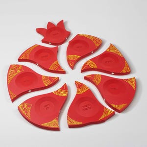 May include: A set of eight red wooden pieces arranged in a circular pattern, each featuring a circular indentation and yellow line art designs. The pieces include Hebrew text engraved within the indentations, forming a segmented display set.