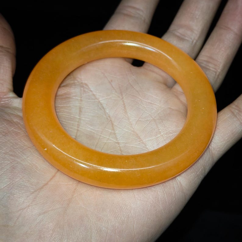 Puede incluir: Un brazalete de color naranja transl&uacute;cido. El brazalete circular es de color s&oacute;lido y parece estar hecho de un material liso y pulido. El brazalete se sostiene en una mano sobre un fondo oscuro.