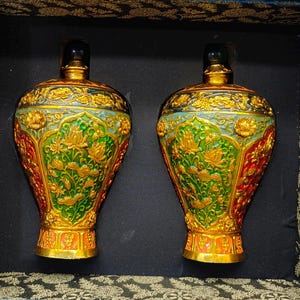 May include: Two matching decorative vases featuring a vibrant green and gold floral relief pattern. Each vase has a rounded body, a narrow neck with a dark stopper, and a flared base. The vases are displayed side-by-side within a dark-lined box.
