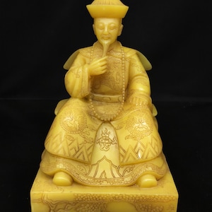 May include: A detailed, yellow-toned statue of a seated figure in traditional attire. The figure wears a hat and holds a beard, with intricate carvings on the clothing and base. The statue is on a rectangular base with additional carvings.