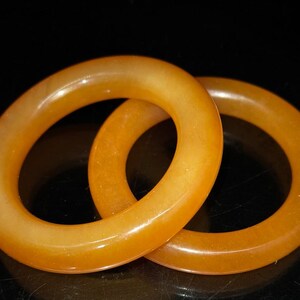 Puede incluir: Dos brazaletes circulares de color &aacute;mbar se muestran sobre un fondo negro. Los brazaletes est&aacute;n hechos de un material transl&uacute;cido, con una superficie lisa y pulida. La luz captura las curvas de los brazaletes, creando reflejos y sombras.