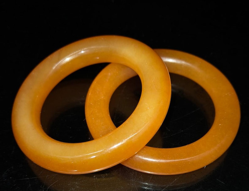 Puede incluir: Dos brazaletes circulares de color &aacute;mbar se muestran sobre un fondo negro. Los brazaletes est&aacute;n hechos de un material transl&uacute;cido, con una superficie lisa y pulida. La luz captura las curvas de los brazaletes, creando reflejos y sombras.