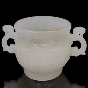 May include: A white, antique-style ceramic vessel with two dragon-shaped handles. The vessel has a wide, open top and a detailed, embossed pattern around the body. The base is a small, circular pedestal. The background is black.