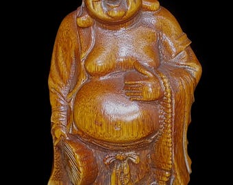 Puede incluir: Una figura de madera tallada de un personaje sonriente y corpulento con una gran barriga, que viste túnicas holgadas. La figura sostiene un pequeño abanico en la mano derecha y un rosario en la mano izquierda, con una veta de madera marrón claro pulida.