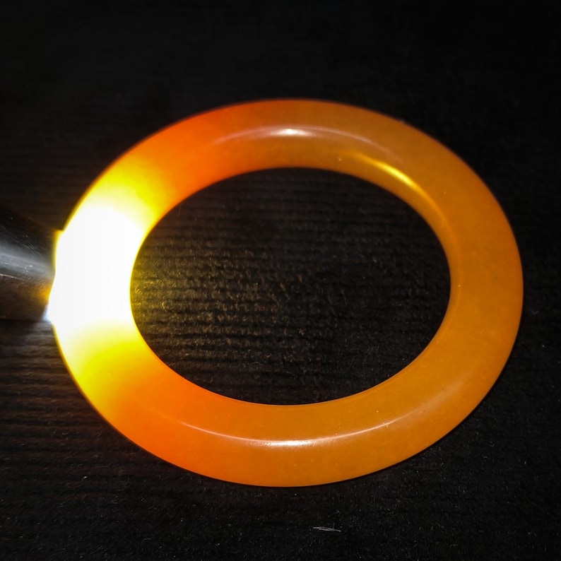 Puede incluir: Un brazalete de color naranja-amarillo transl&uacute;cido. El brazalete circular est&aacute; iluminado desde un lado, creando un brillo brillante. La superficie lisa y pulida refleja la luz, resaltando su color y forma. El fondo es oscuro.