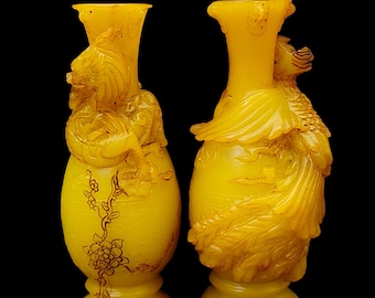 Puede incluir: Dos jarrones amarillos translúcidos con intrincadas tallas en relieve. El jarrón izquierdo muestra una figura de dragón y un diseño de rama floral. El derecho presenta una figura de fénix con plumaje detallado. Ambos tienen borde y base acampanados.