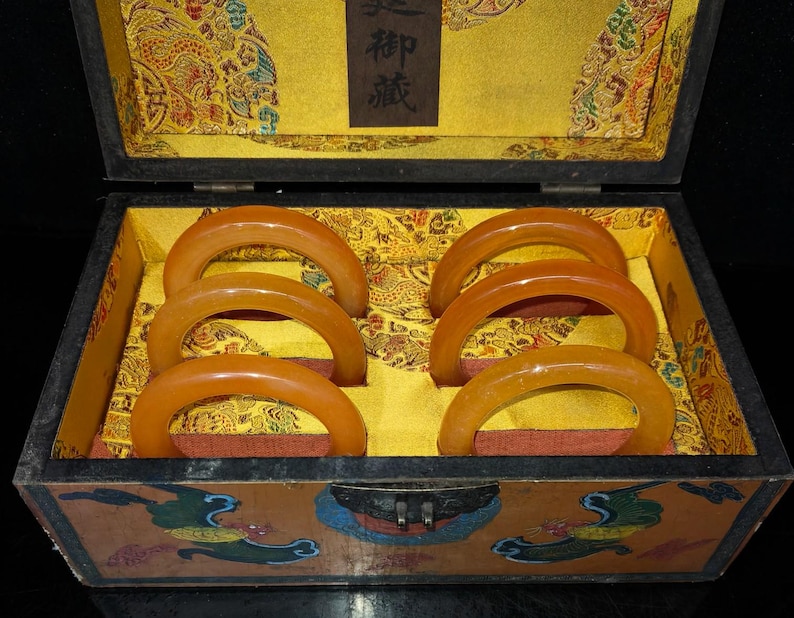 Puede incluir: Caja de madera abierta con seis brazaletes de color &aacute;mbar. El interior de la caja est&aacute; forrado con tela amarilla y una base roja. El exterior de la caja presenta dise&ntilde;os pintados, mostrando los brazaletes.