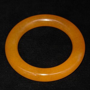 Puede incluir: Un brazalete liso de color &aacute;mbar. El brazalete circular tiene un acabado pulido y un tono dorado c&aacute;lido. La joya est&aacute; colocada sobre un fondo negro, lo que resalta su color y forma. Este accesorio es adecuado para el uso diario.