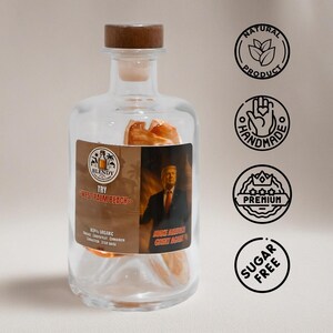 May include: A clear glass bottle with a wooden stopper, containing orange-colored fruit slices. The label reads "BLENDY TRY WEST PALM BEACH" with text "MAKE AMERICA GREAT AGAIN". Additional circular labels indicate "NATURAL PRODUCT", "HANDMADE", "PREMIUM", and "SUGAR FREE".