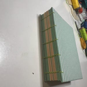 May include: A hand-bound book featuring a light green and white striped fabric cover. The spine is exposed, showcasing alternating light green and peach-colored paper signatures held together by visible lime green thread stitching.