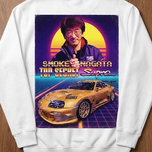 以下が含まれることがあります： ゴールドのトヨタスープラと「Smokey Nagata Top Secret Supra」の文字がプリントされた白いスウェットシャツ。デザインには男性の肖像画とレトロなスタイルの背景が含まれています。