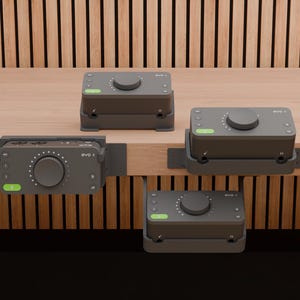 Peut inclure: Quatre interfaces audio grises avec des accents noirs et des voyants verts sont disposées sur une surface en bois. Les interfaces ont un bouton de commande circulaire et le texte "EVO 4". L'arrière-plan présente des lattes de bois verticales.